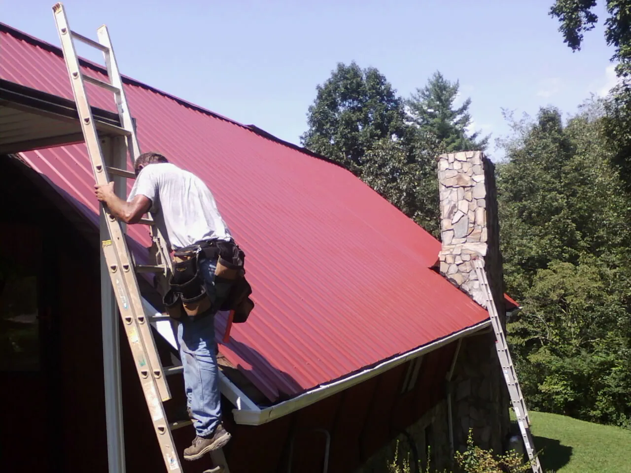 Roofer on a ladder beside a red metal roof and stone chimney — Soffit & Fascia Repair by Roofing Brothers in Donegal